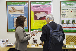 木材プレス成型品を手に取りながら研究成果を説明している研究者と熱心に解説を聞いている来場者